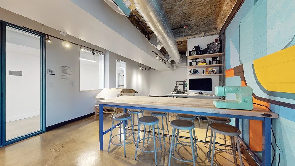 Workshop with long table and stools, sewing machine, and colorful mural on the wall.