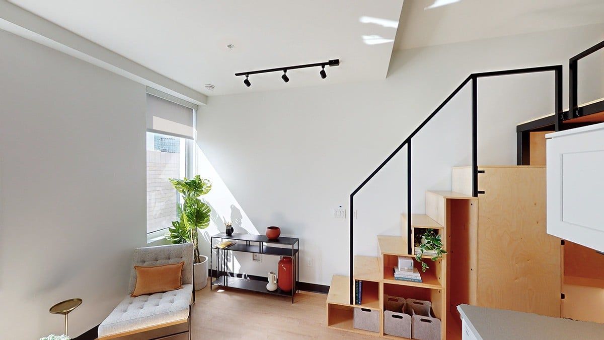 Living room with a loft, stairs, and shelves. Natural light from a window and neutral colors.