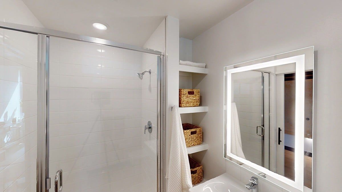 Bathroom with a glass shower, lit mirror, white walls, and woven storage baskets on shelves.