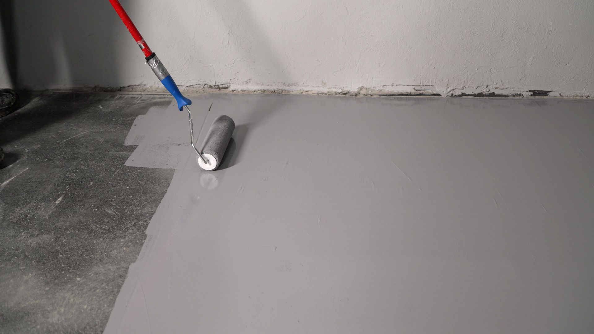 A person is rolling gray paint onto a concrete floor with a roller and extended handle.