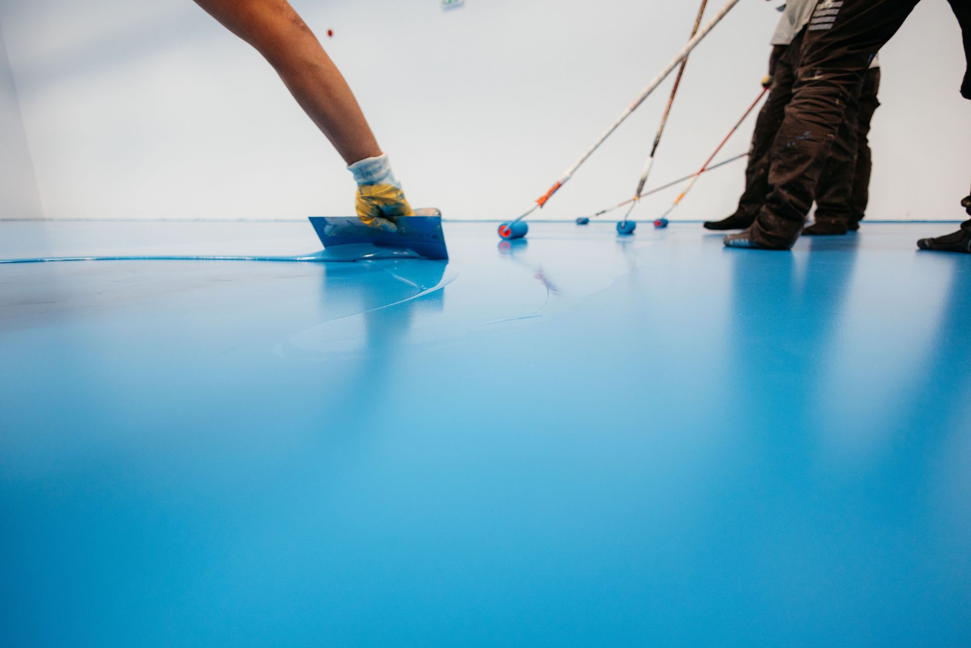 Workers applying blue epoxy coating to a floor with rollers and a trowel in a white room.