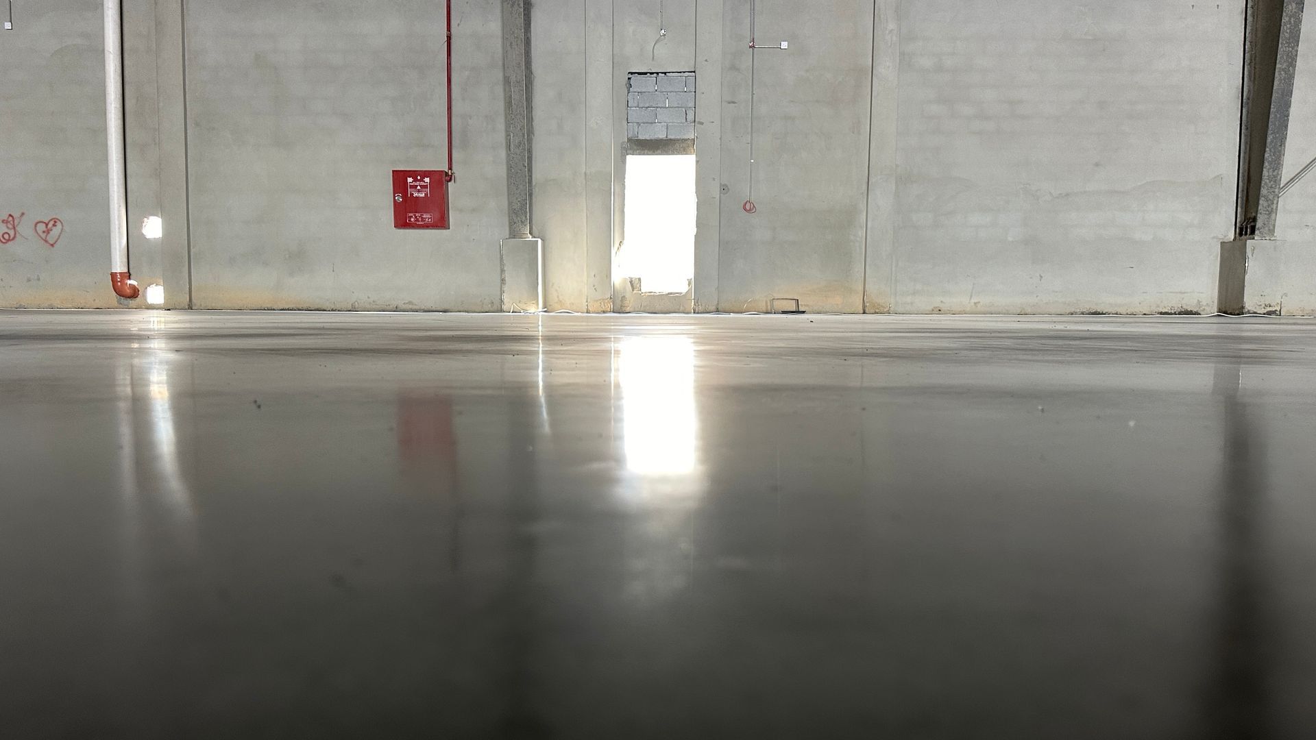 A shiny, gray concrete floor in a large, empty industrial building with a bright doorway and fire equipment.