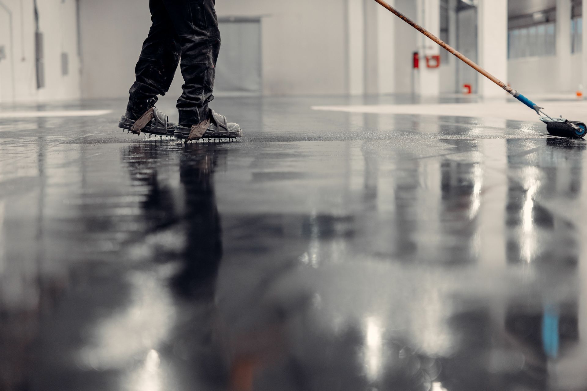 Worker wearing spiked shoes rolling epoxy coating on a glossy industrial floor.