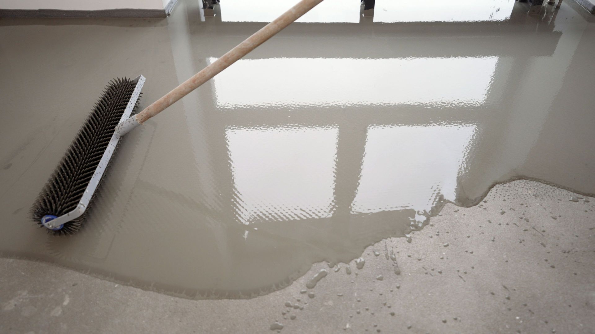 Freshly poured, wet, gray concrete floor being smoothed with a long-handled brush, window reflection visible.