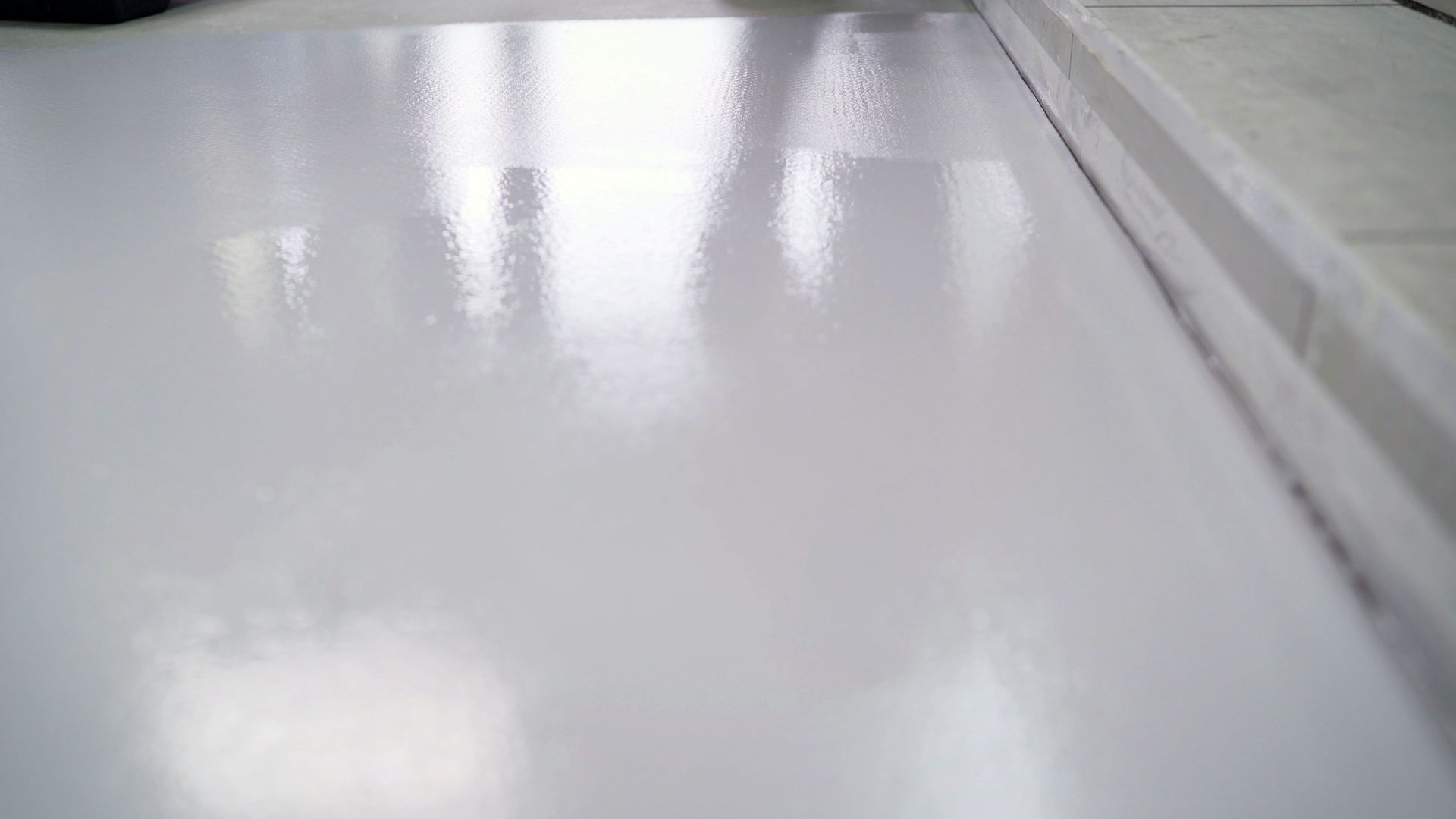 Shiny, light gray painted floor reflecting light. Edge of a room with a white border visible.