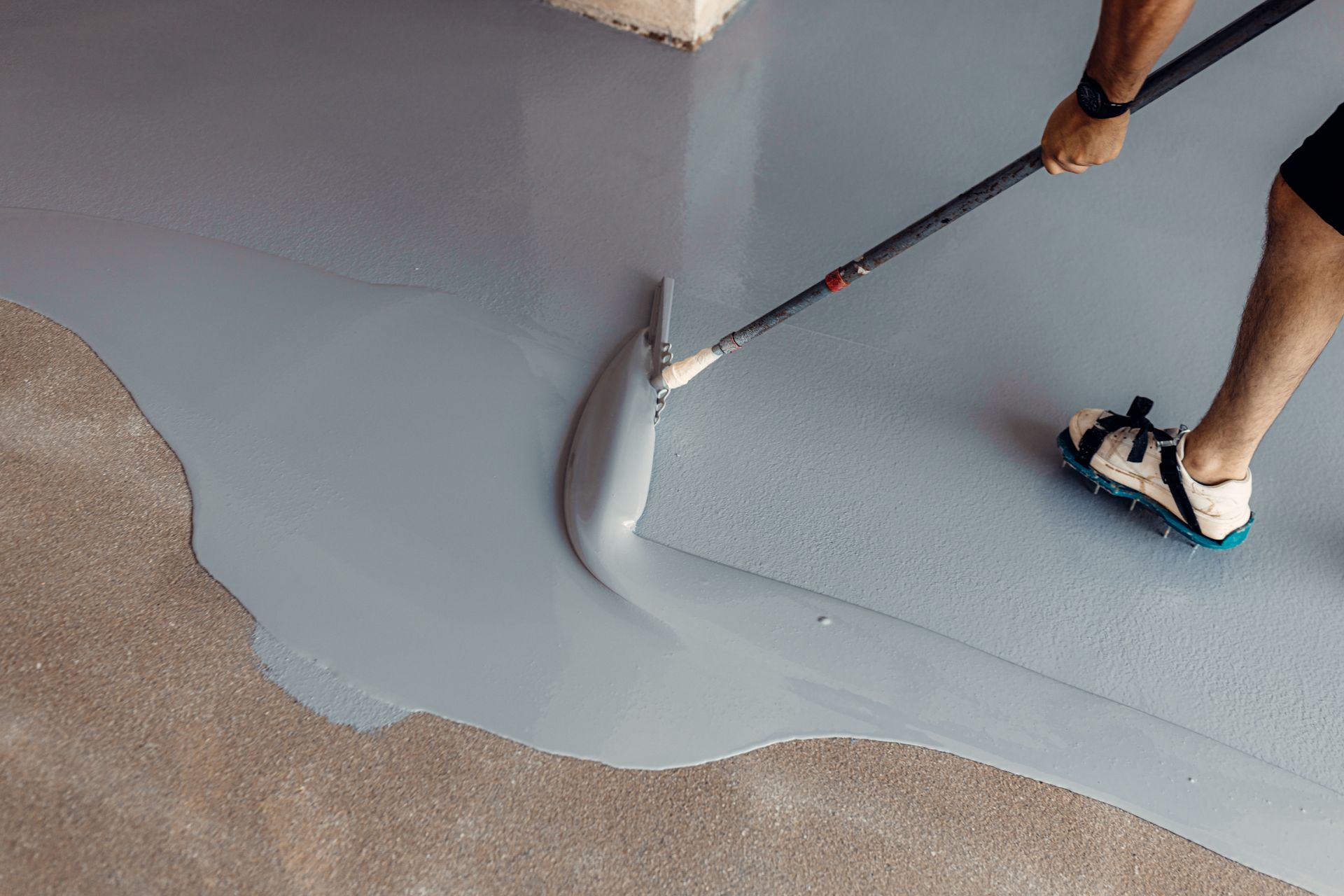 Person using a squeegee to apply gray epoxy floor coating in a garage.