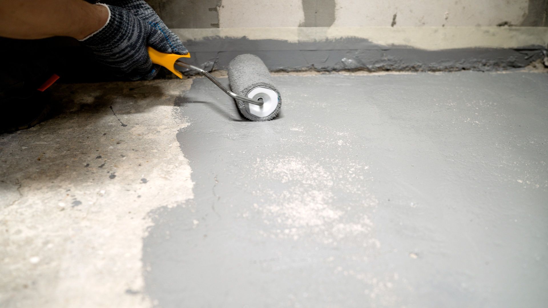 A person is using a paint roller to apply gray sealant to a concrete floor.