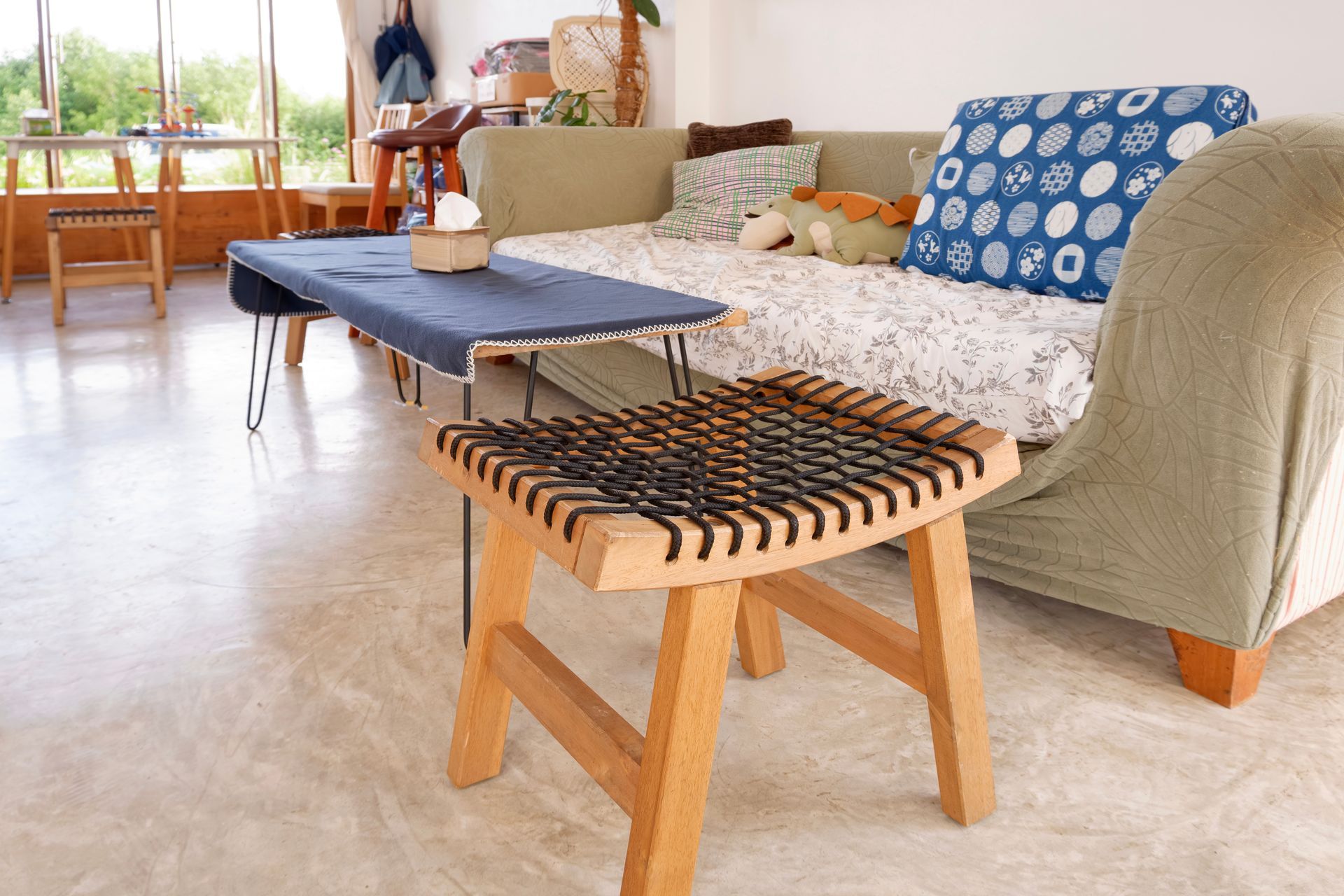 Living room with a wooden stool, couch, and a blue-covered table. Natural light floods the room.