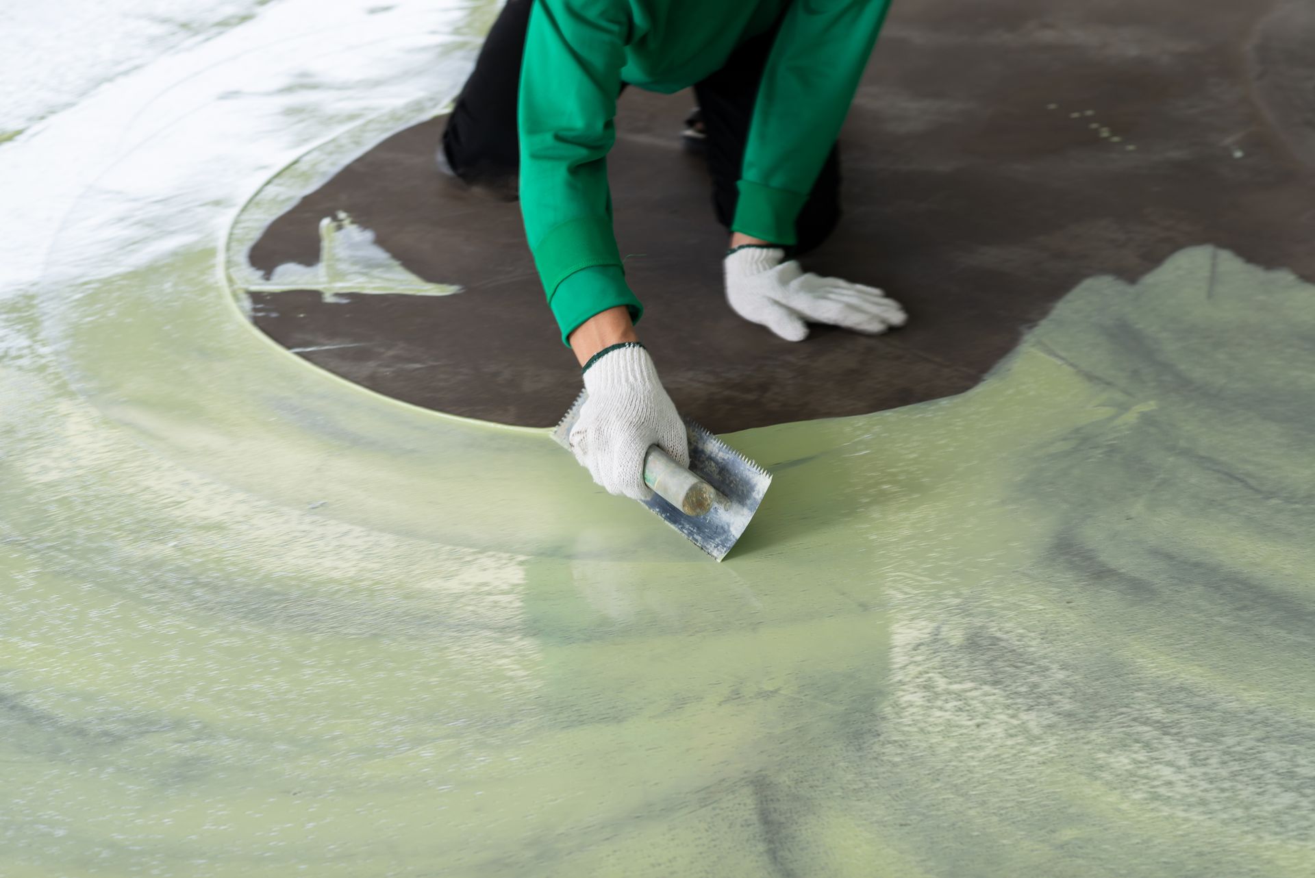 Person kneeling, applying light green sealant to a concrete floor with a trowel, wearing gloves and a green shirt.