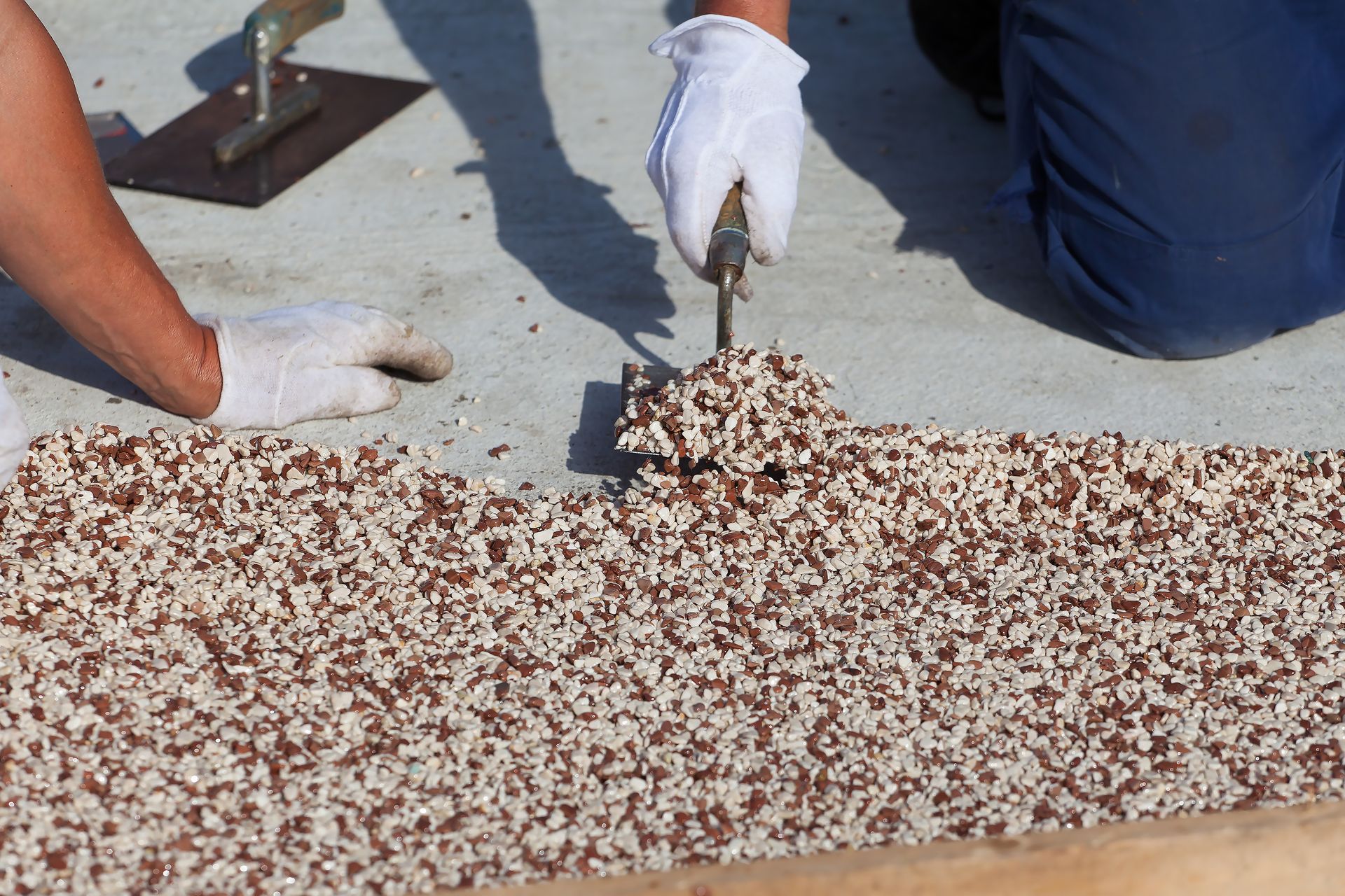 Hands in gloves spreading aggregate material on a surface with a trowel.