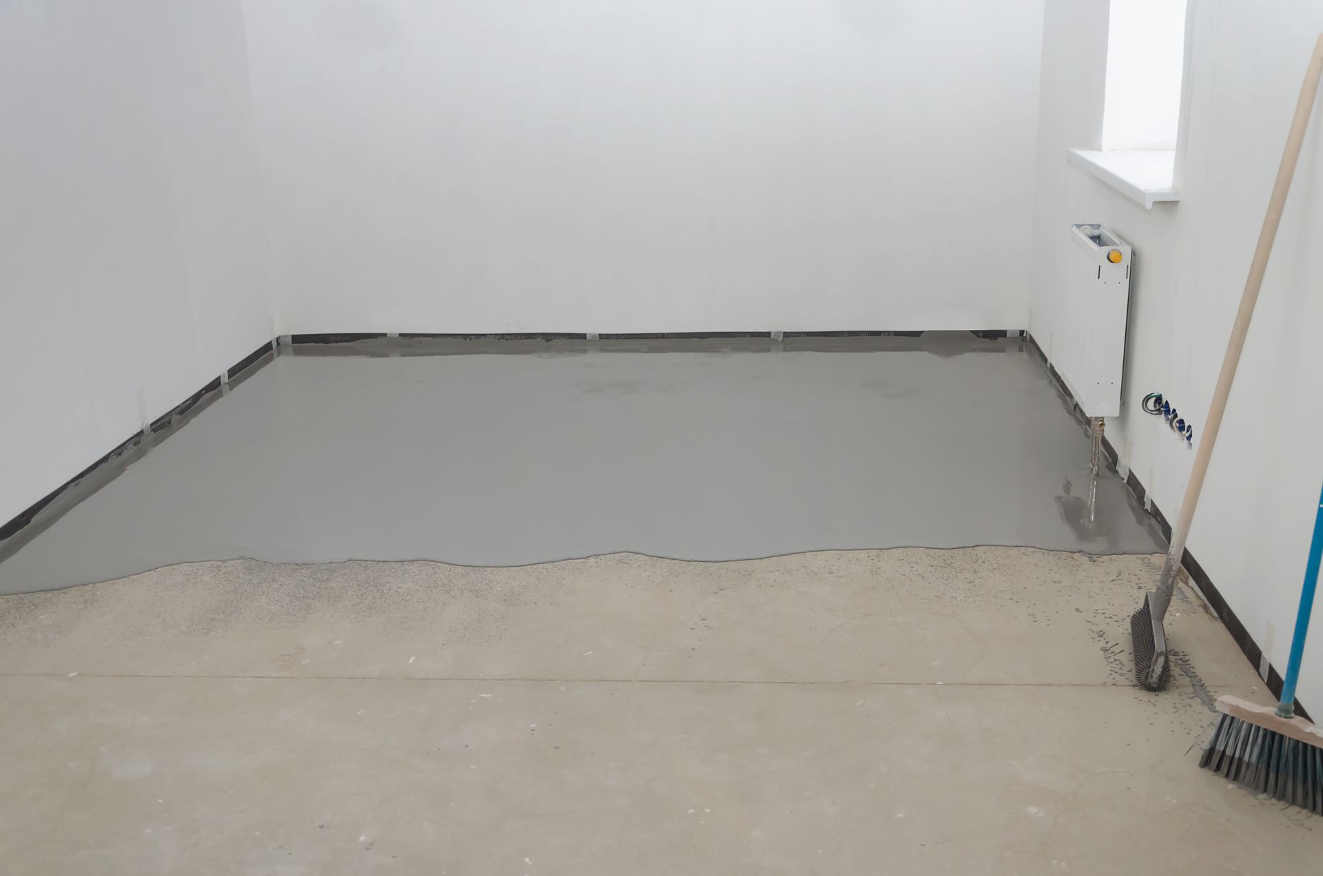 Room with wet, gray floor screed, ready for flooring. Broom leans against the wall.