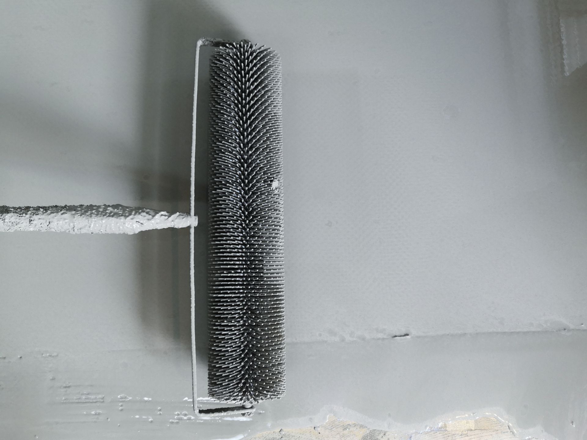 A spiked roller tool is used to remove air bubbles from a freshly applied grey coating on a wall.