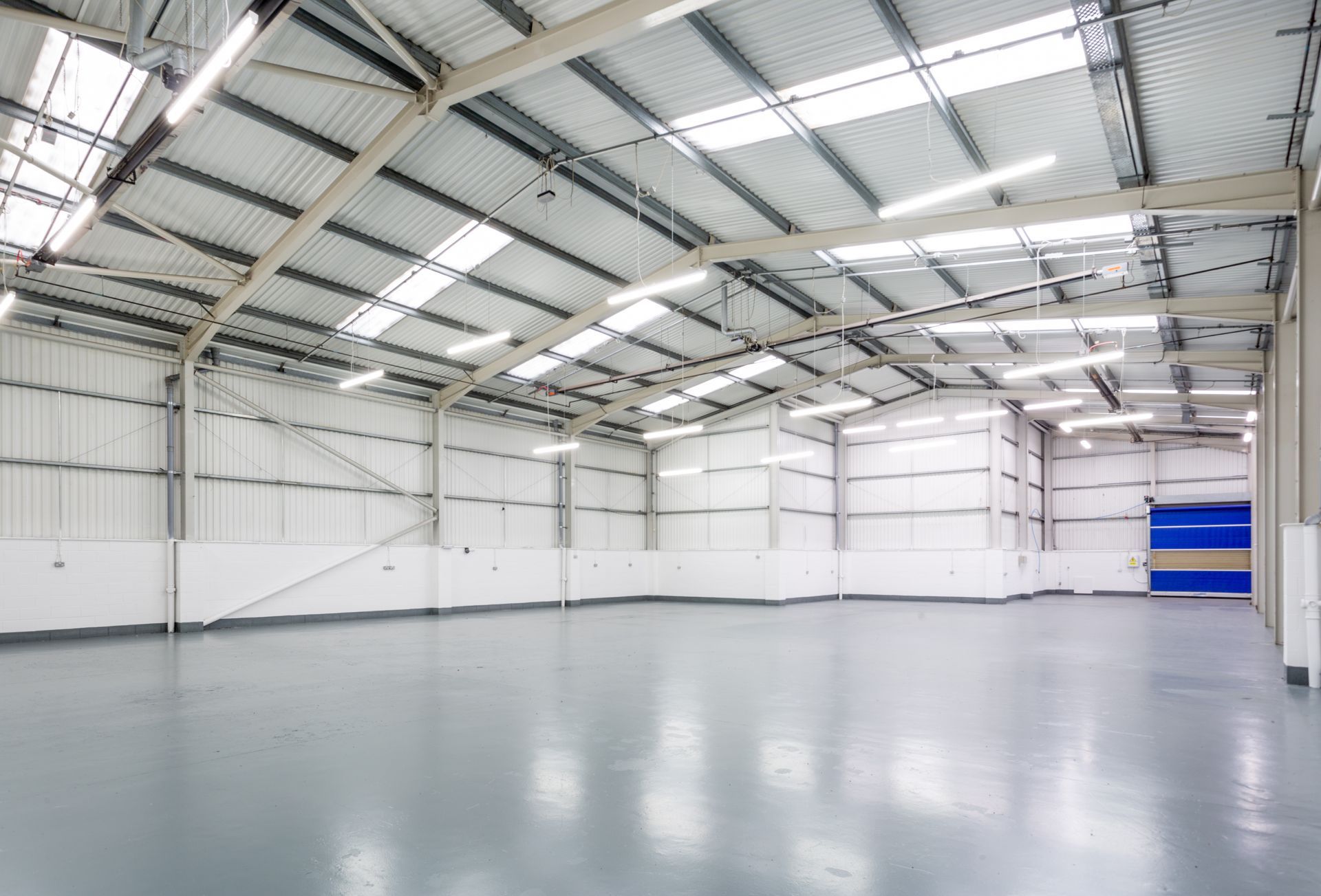 Empty warehouse interior with a gray floor and corrugated metal ceiling, lit by overhead lights and skylights.