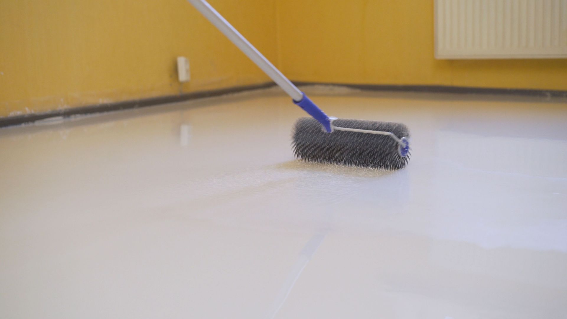 A person using a spiked roller to smooth out a light-colored floor coating in a room with yellow walls.