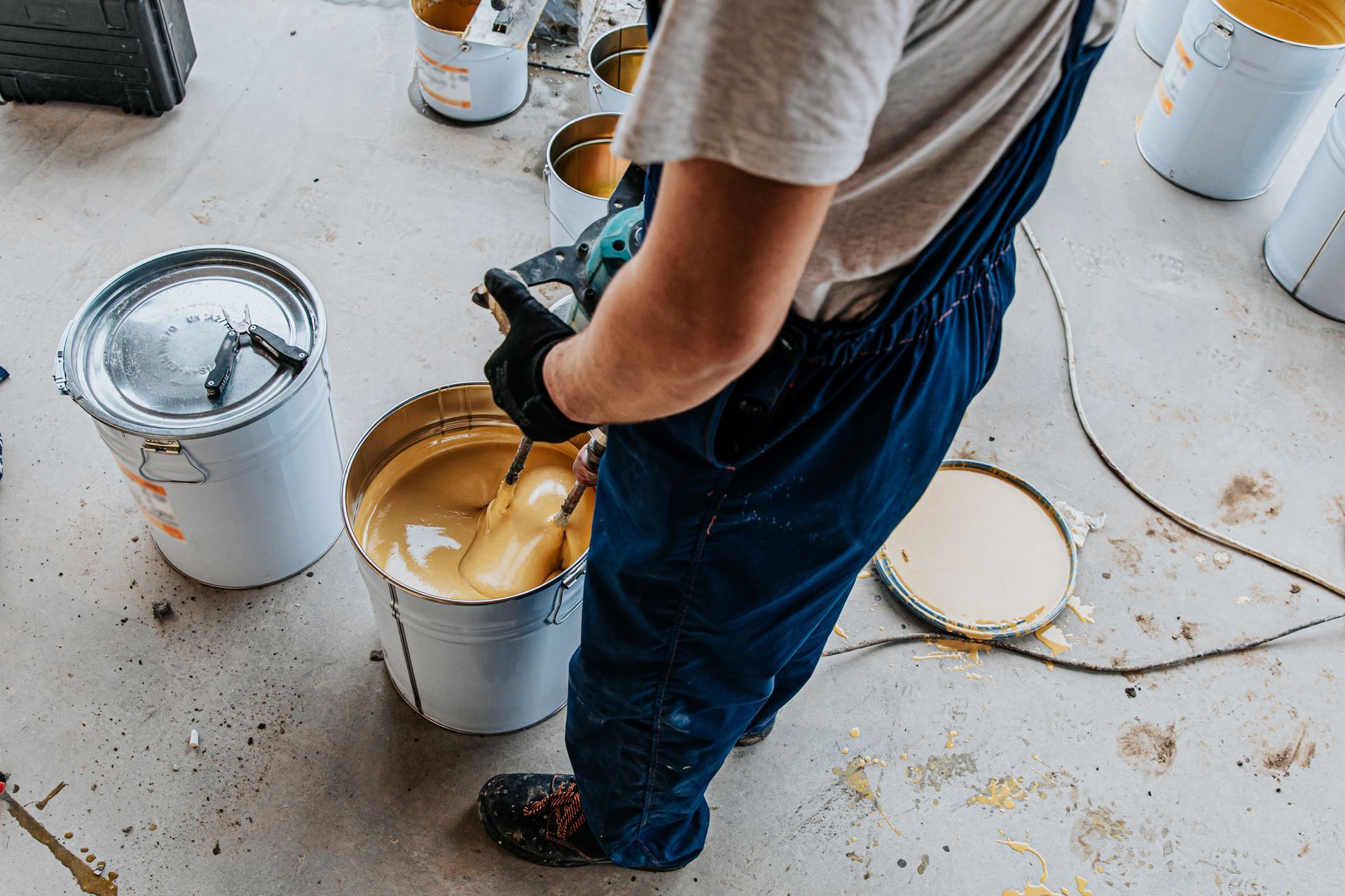 Person mixing paint in a bucket with a power mixer; several paint cans on floor.