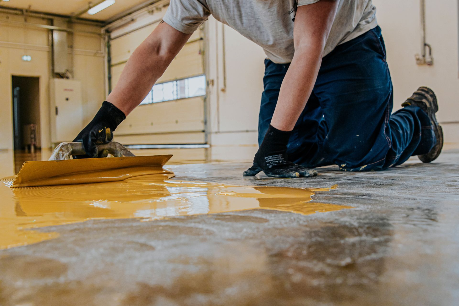 Person kneeling, applying yellow epoxy floor coating with a trowel in a garage.