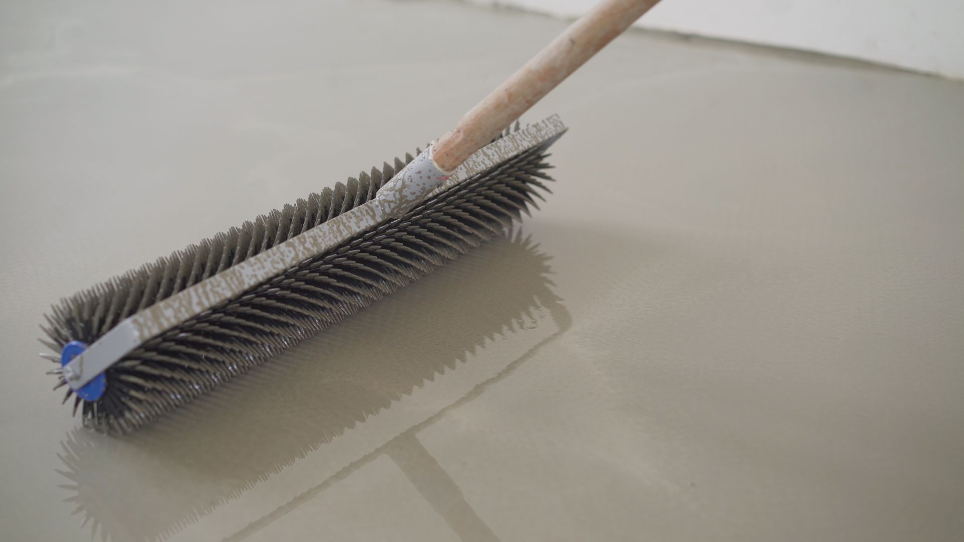 Long-handled brush smoothing wet, gray floor coating, reflecting light.
