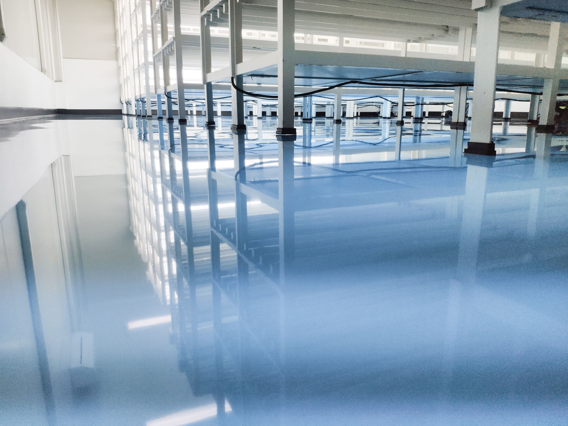 Blue, reflective industrial epoxy floor; white pillars and shelving in a large factory or warehouse setting.