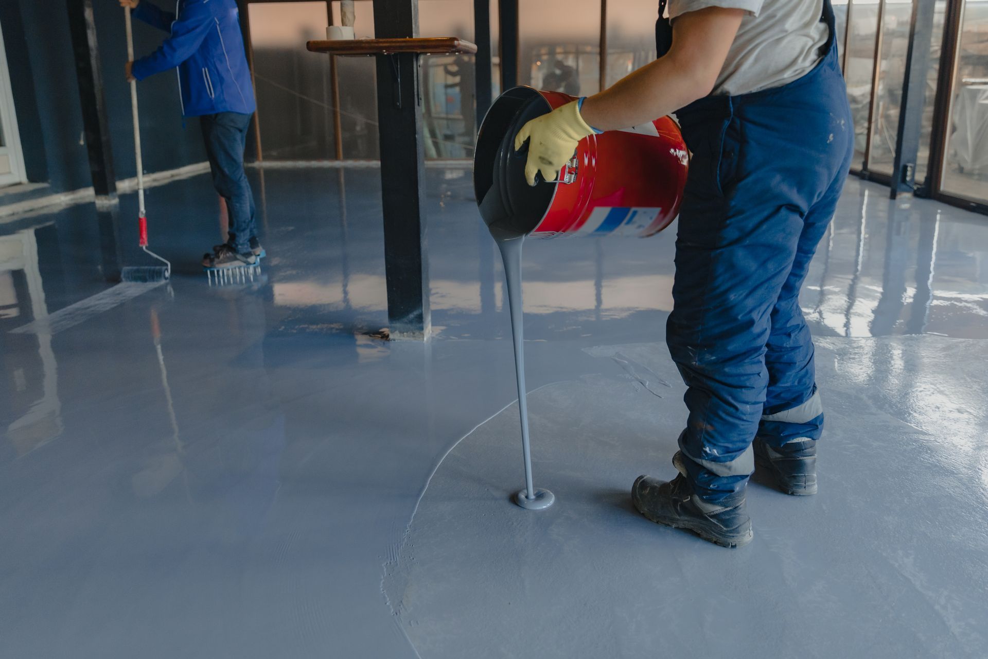 Person pouring grey epoxy from a bucket onto a floor, another rolling it out, indoor setting.