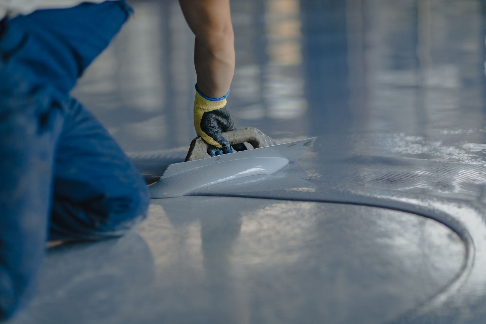 Person using a trowel to smooth gray epoxy floor coating in a large indoor space.