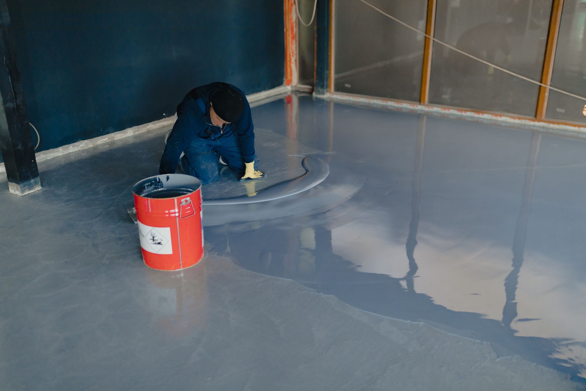 Man kneeling, applying gray epoxy floor coating in a room. Red bucket nearby.