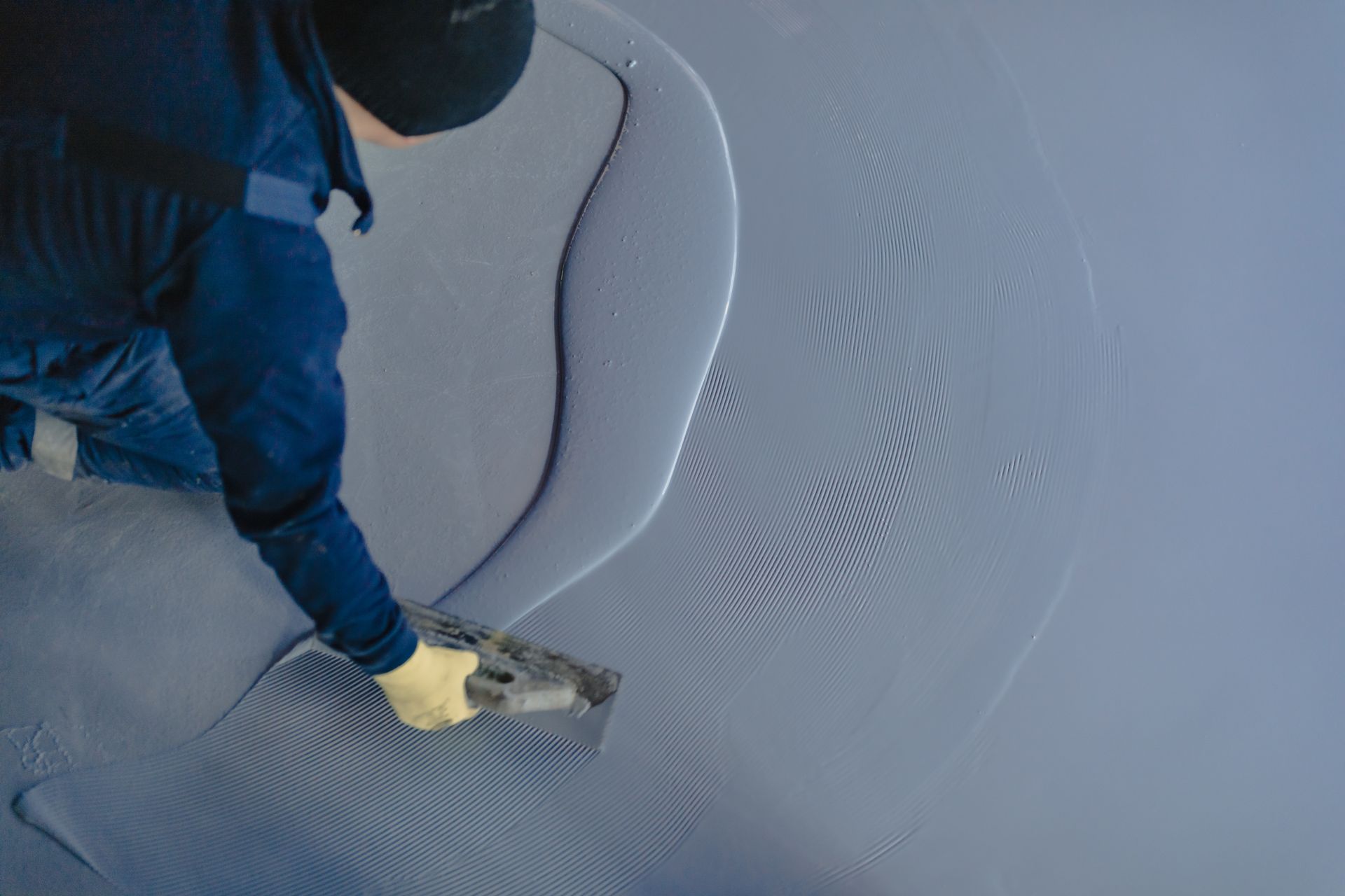 Person in blue work clothes spreading blue sealant on floor with a trowel.