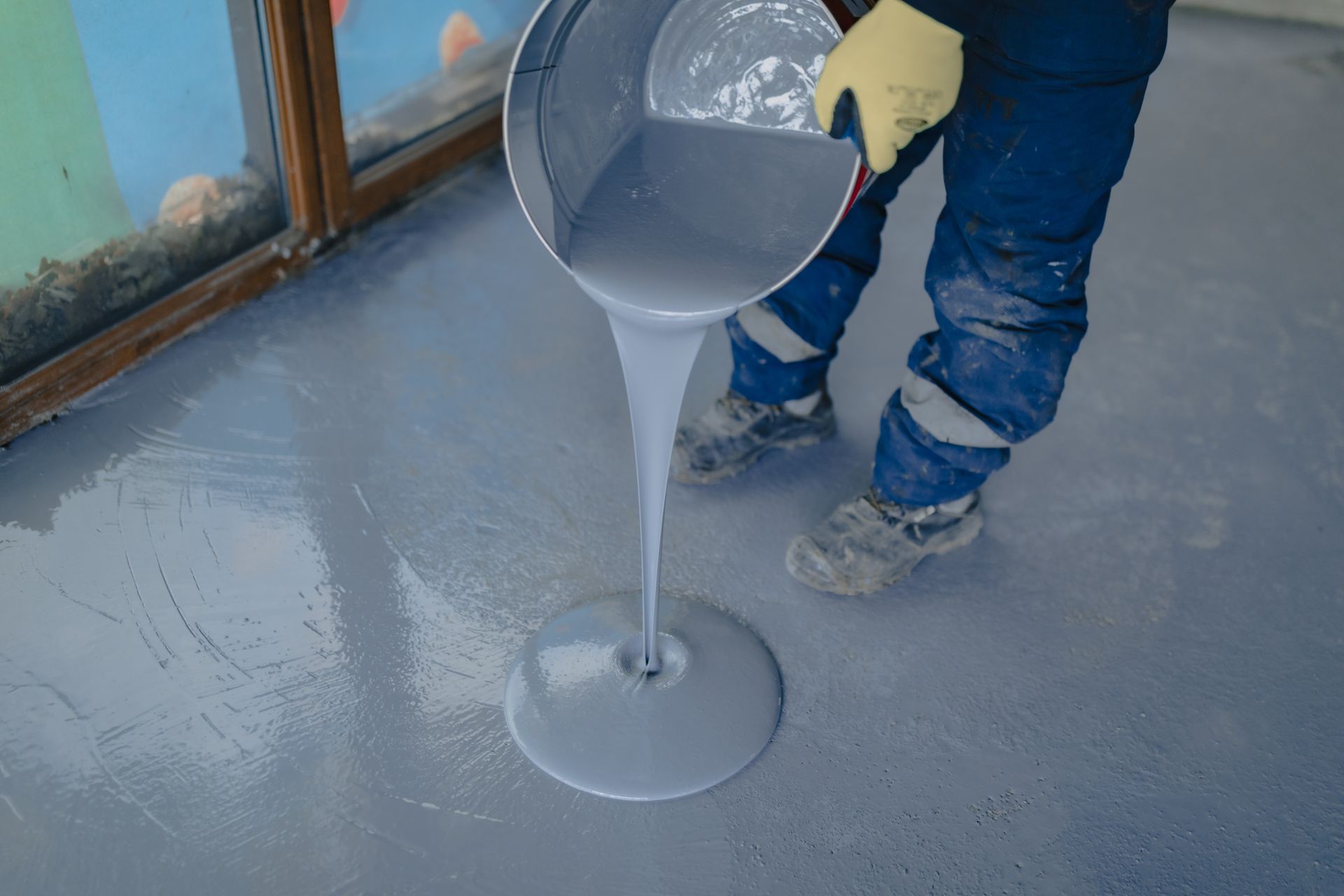 A person pouring gray epoxy onto a gray floor, wearing blue pants and gloves, inside a building.