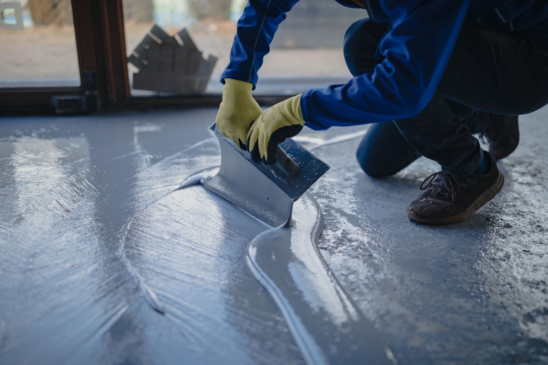 Person applying gray epoxy flooring with a trowel, wearing gloves, indoors.