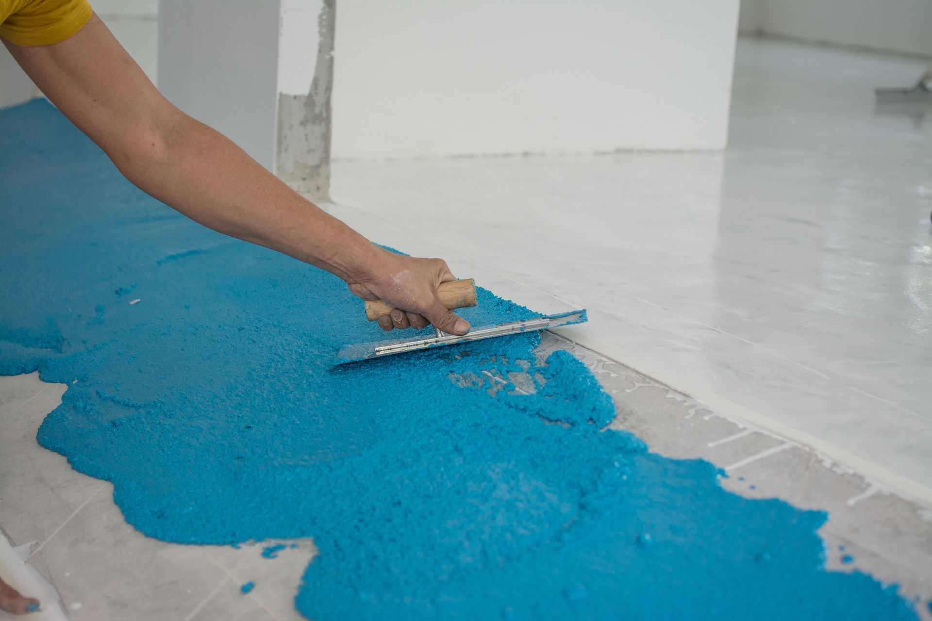 Person smoothing bright blue material onto a light-colored floor with a trowel in a room.