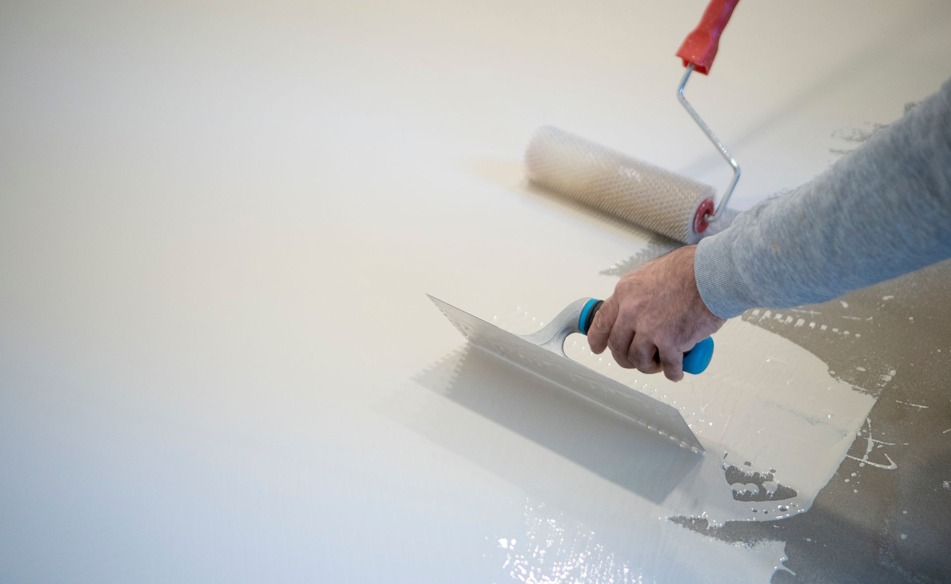 Person smoothing wet white floor coating with a trowel, paint roller in background.