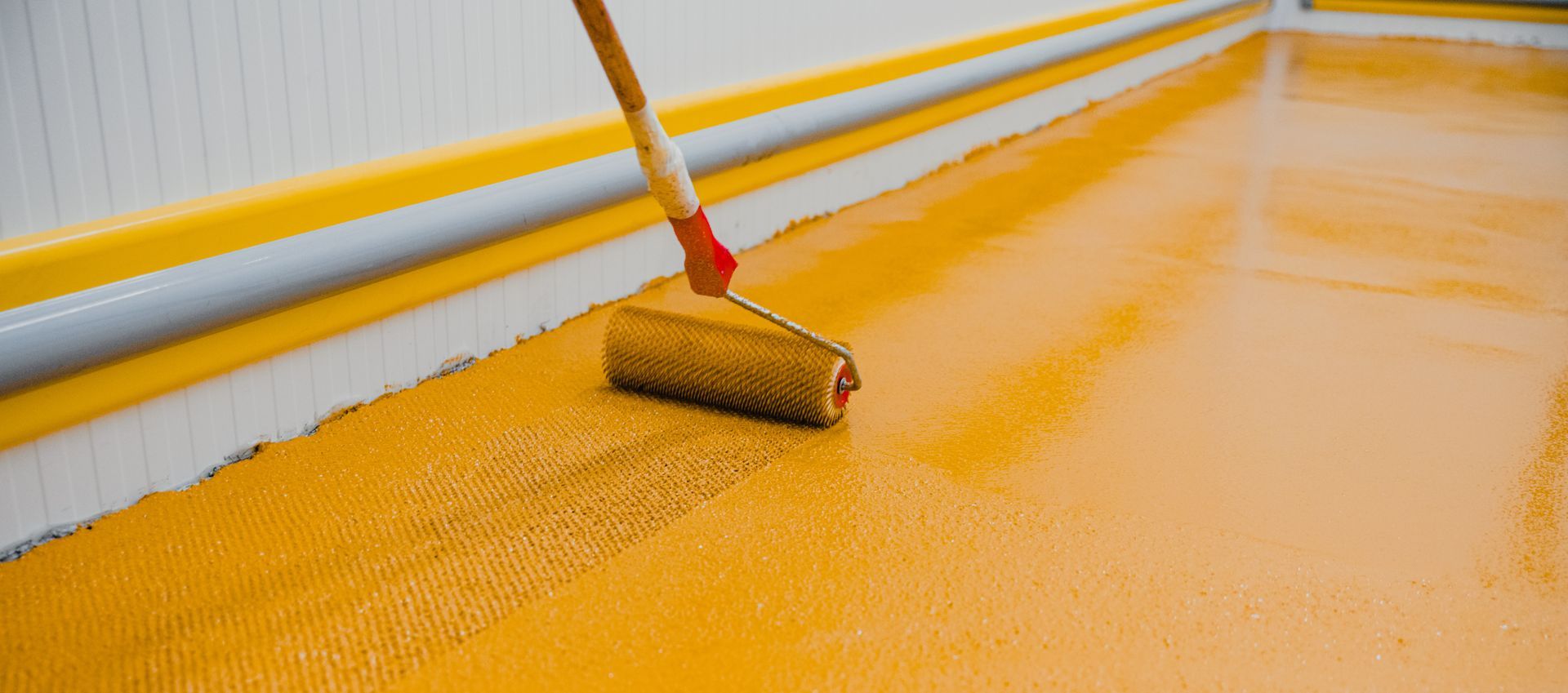 A person using a paint roller applies yellow epoxy flooring to a concrete surface.
