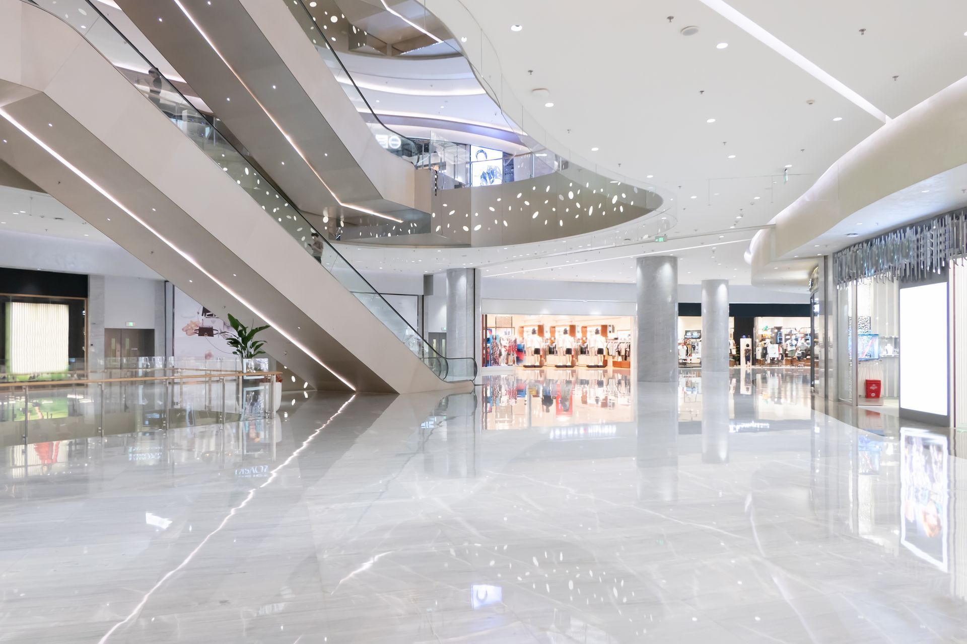 Brightly lit modern shopping mall interior with escalators, marble floors, and storefronts.