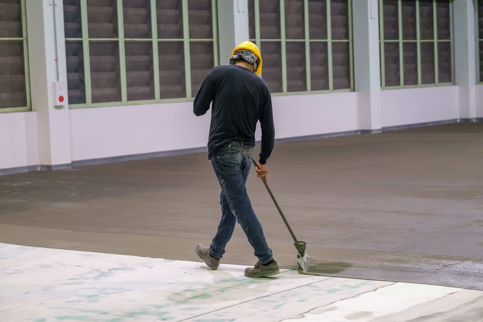 Construction worker, yellow hard hat, applying sealant to a concrete floor with a roller. Interior setting.