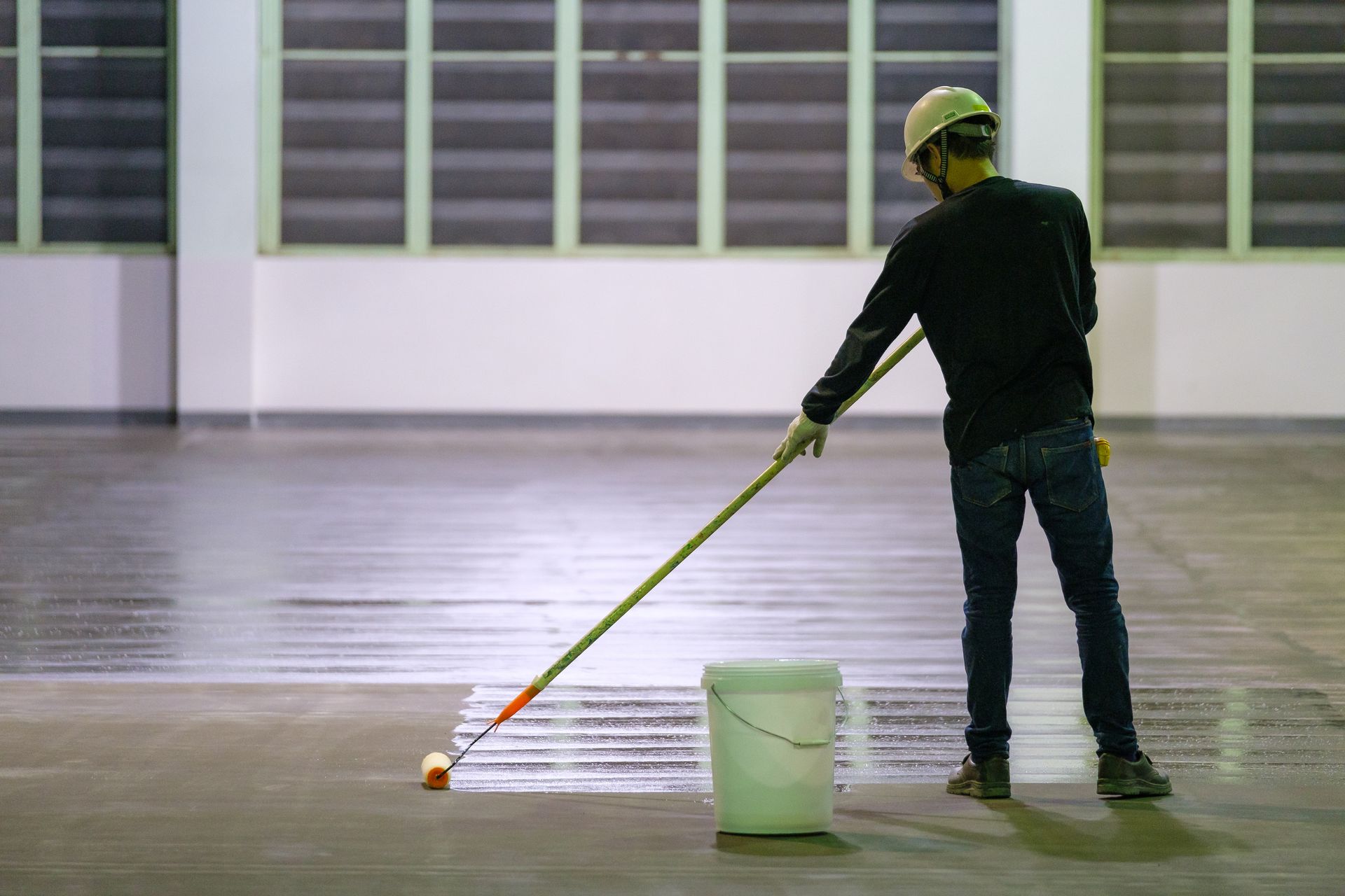 Person in a hard hat and gloves applying coating to a floor with a roller. White bucket nearby.