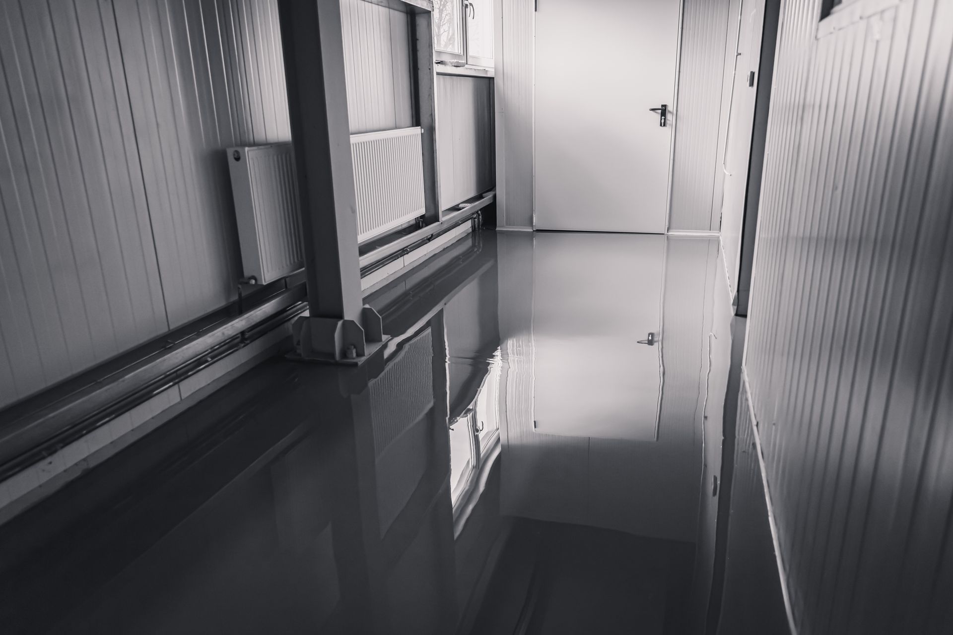 Gray, reflective floor in a narrow hallway with white panel walls and a closed white door at the end.