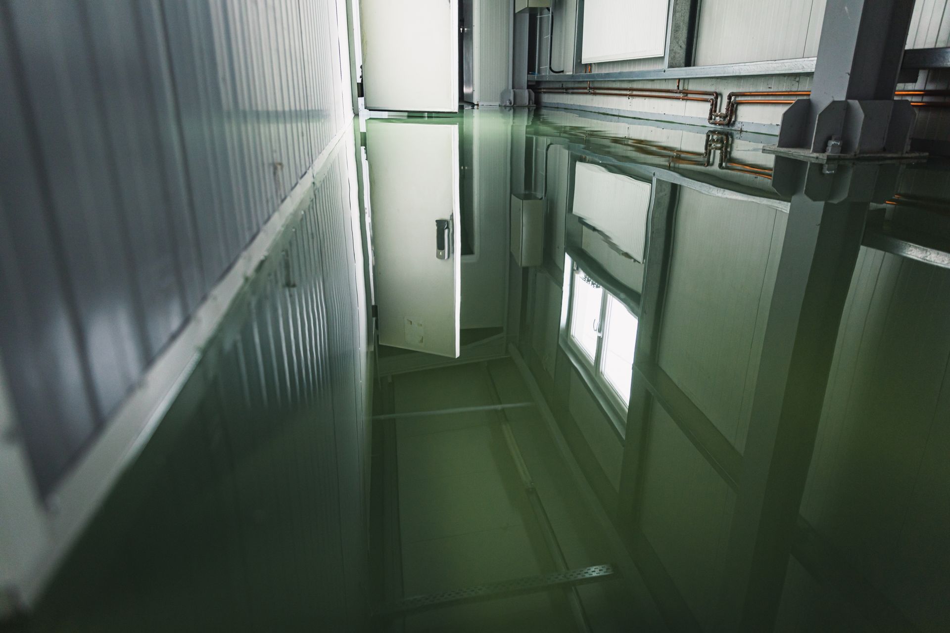 Flooded industrial interior, water reflects a window and door. Green-tinted water fills the room.