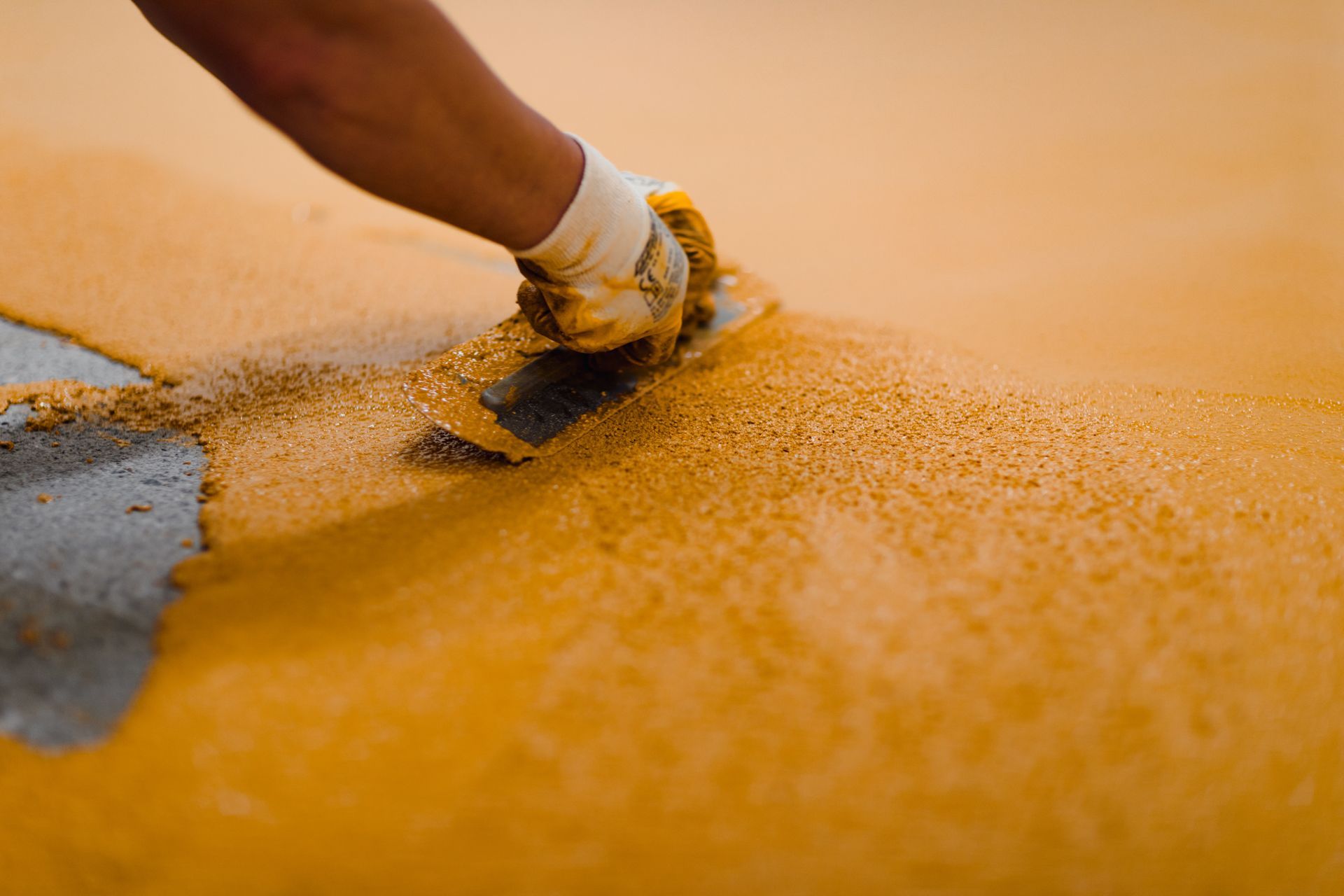 A gloved hand uses a trowel to smooth yellow epoxy over a concrete floor.