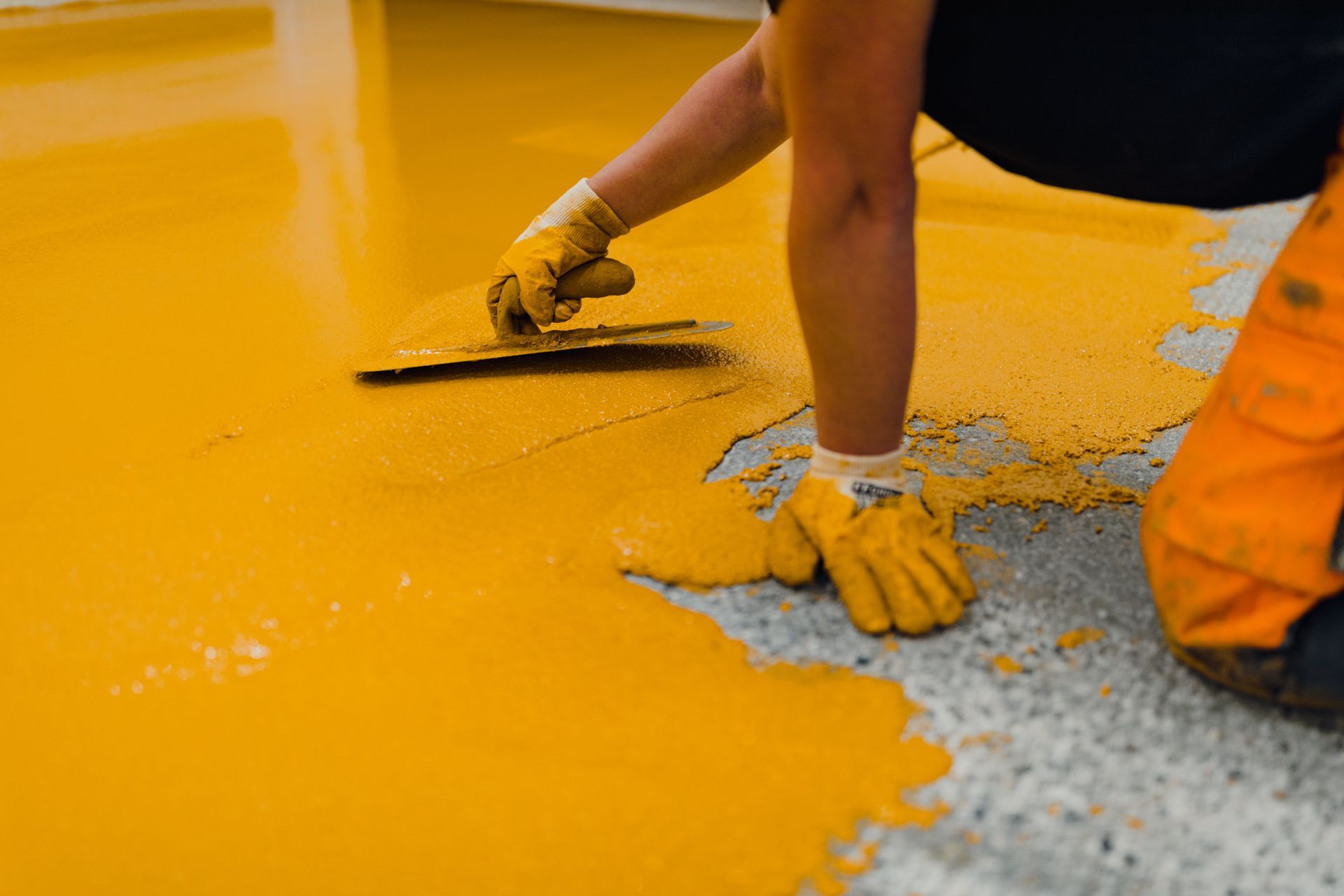 Person spreading yellow epoxy flooring with a trowel, wearing gloves, in a concrete area.