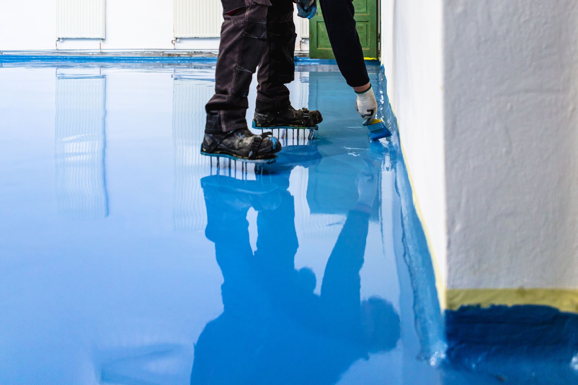 Person applying blue epoxy floor coating with a brush, reflected in the wet surface.