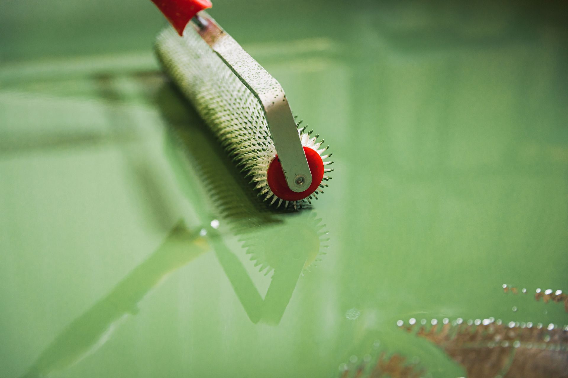 A spiked roller tool in use on a green, liquid surface, creating texture.