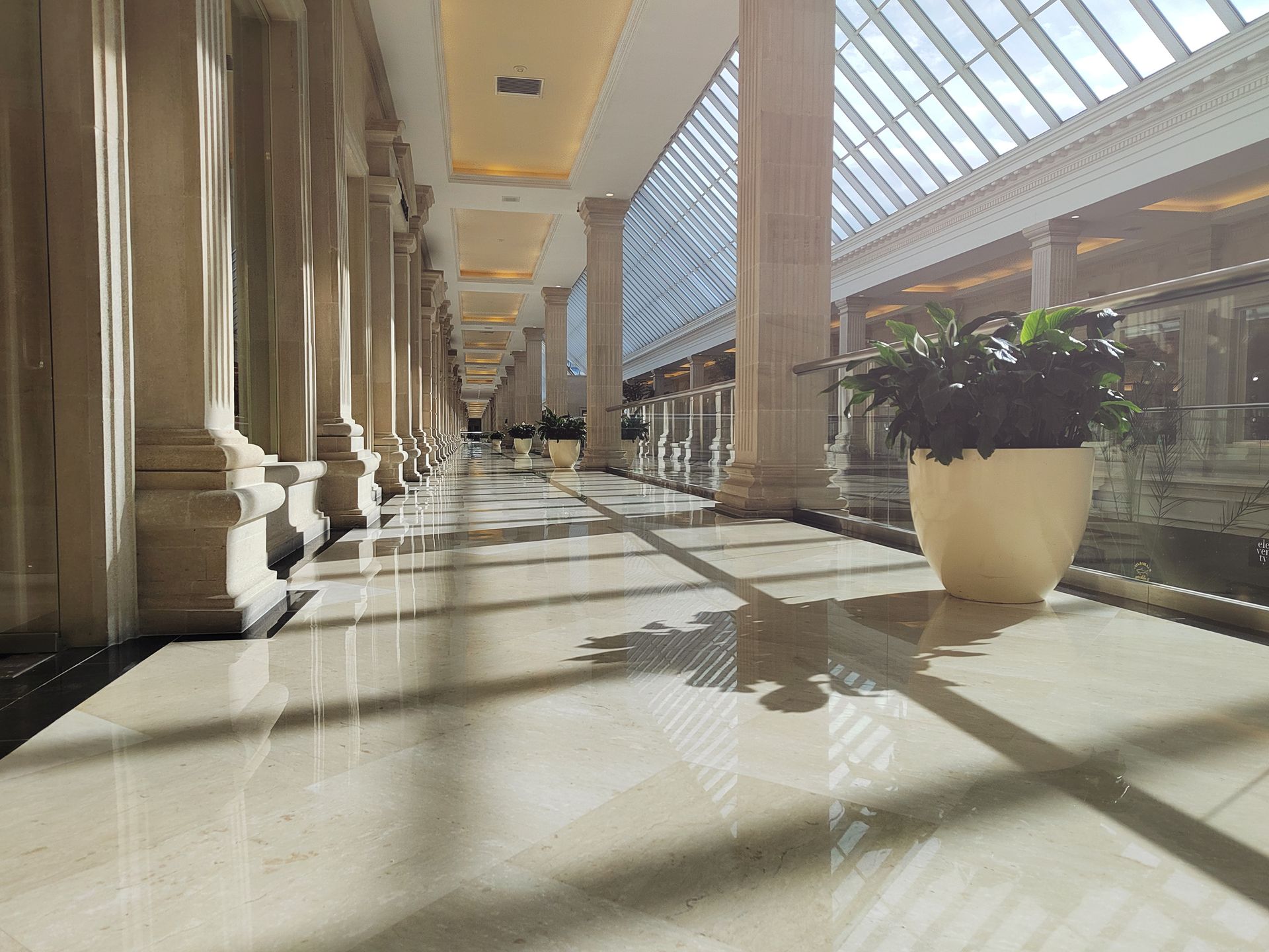 Long hallway with columns, sunlight from skylight, potted plant.