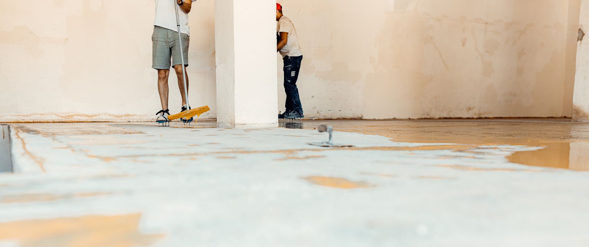 Two people stand in a room, one on a skateboard, water on the floor, unfinished construction.