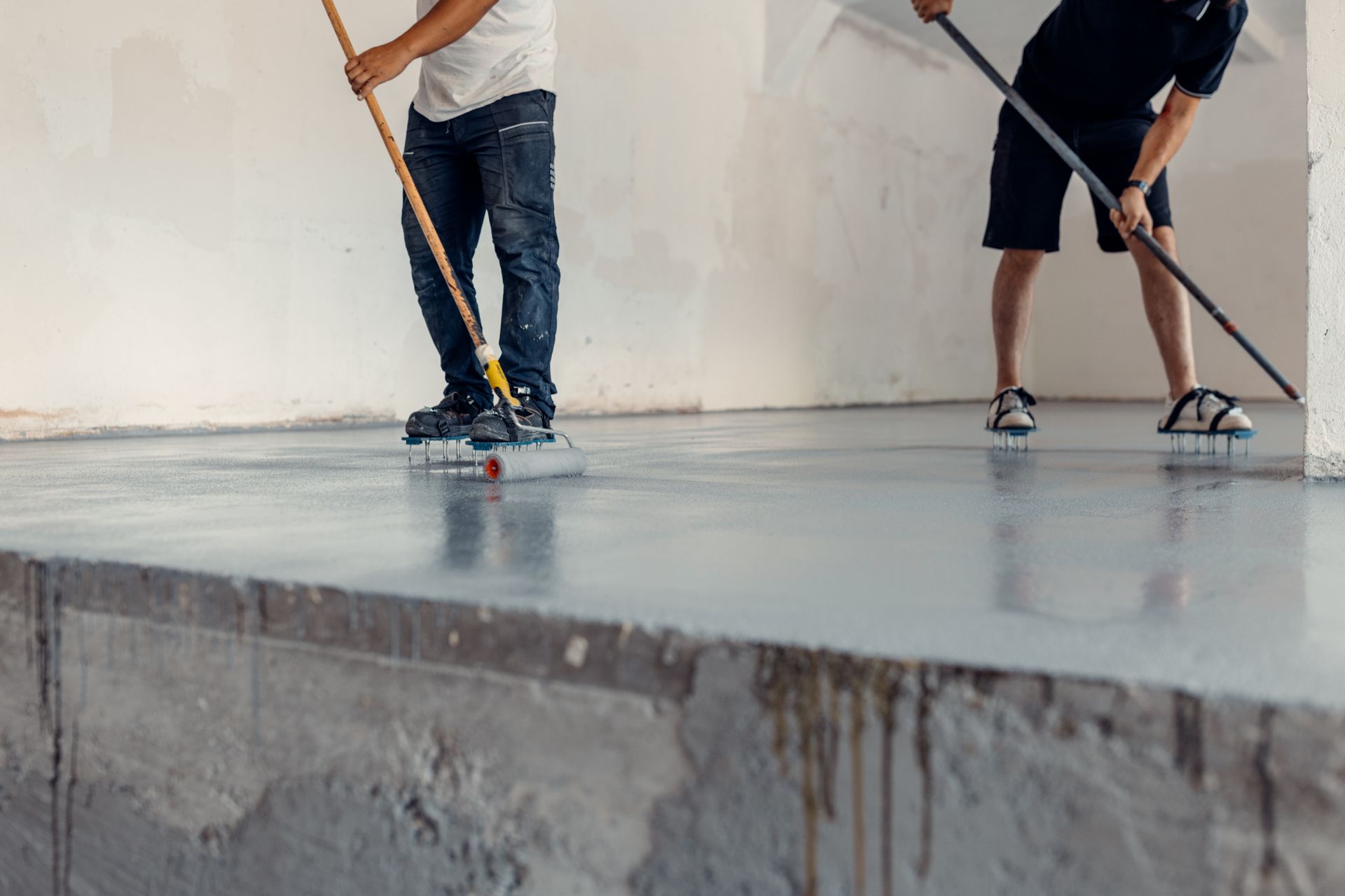 Two people applying a glossy coating to a concrete floor in a room.