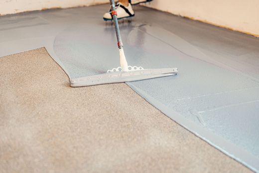 Person applying grey epoxy coating to a concrete floor with a squeegee.