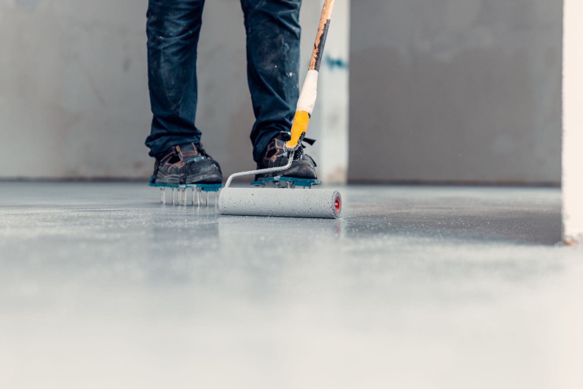Person in jeans paints a floor with a roller. The floor and the person's shoes are blue.