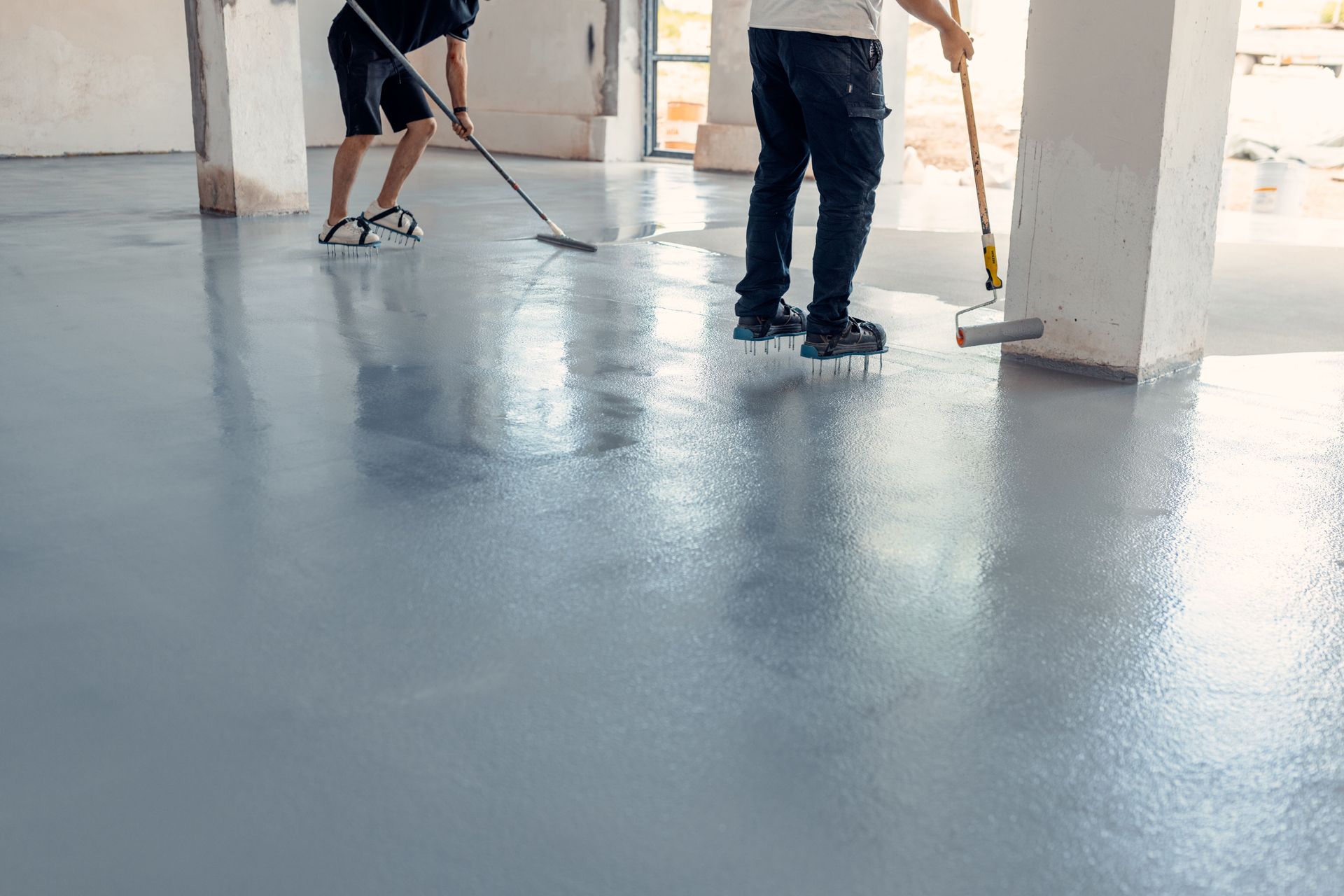 Workers applying epoxy flooring in a light gray room, using rollers and a squeegee.