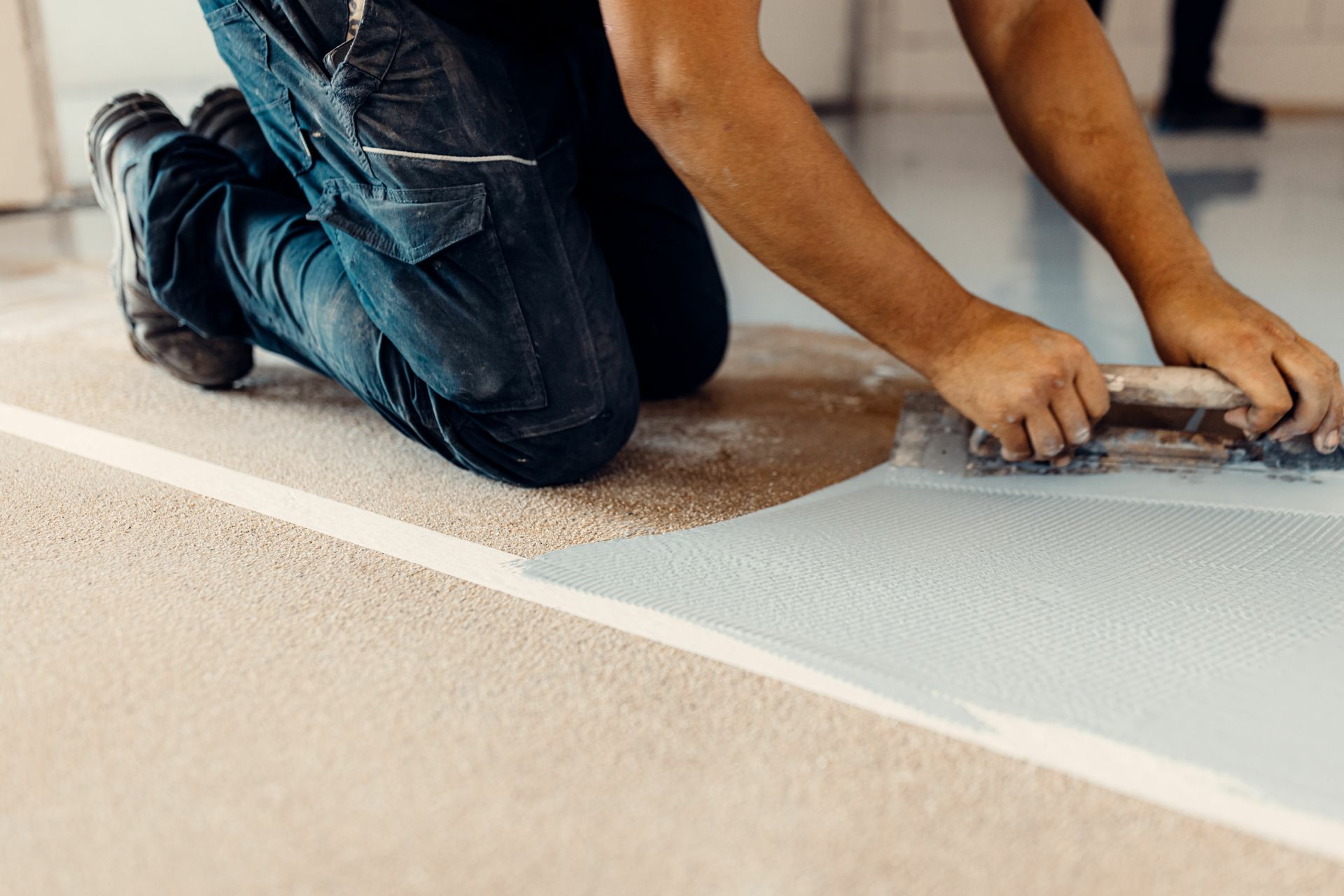 Person kneeling, applying epoxy floor coating with a trowel. White and tan flooring with a blue section.