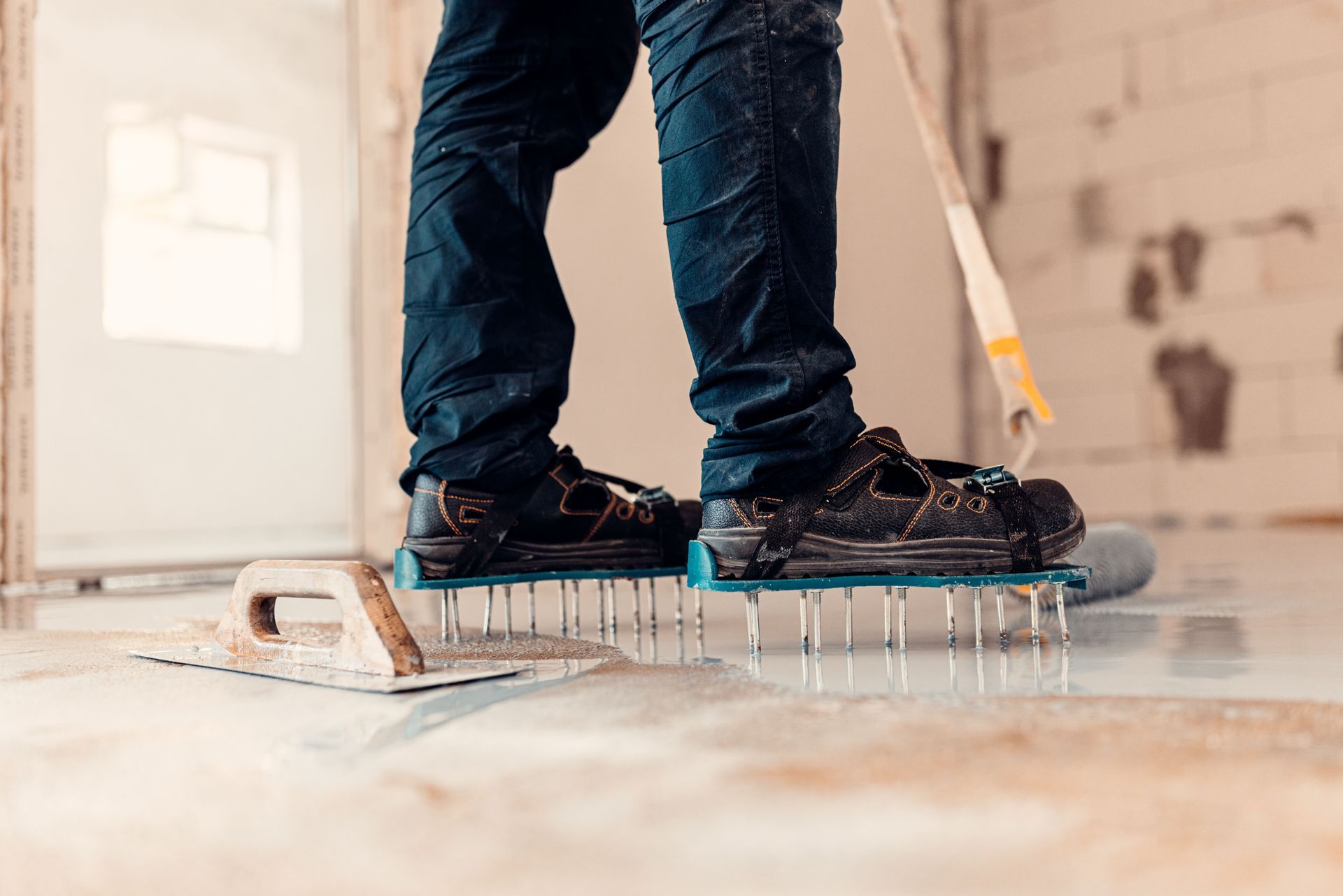 Person rolling concrete, standing on spiked shoes, next to a trowel and roller, indoors.