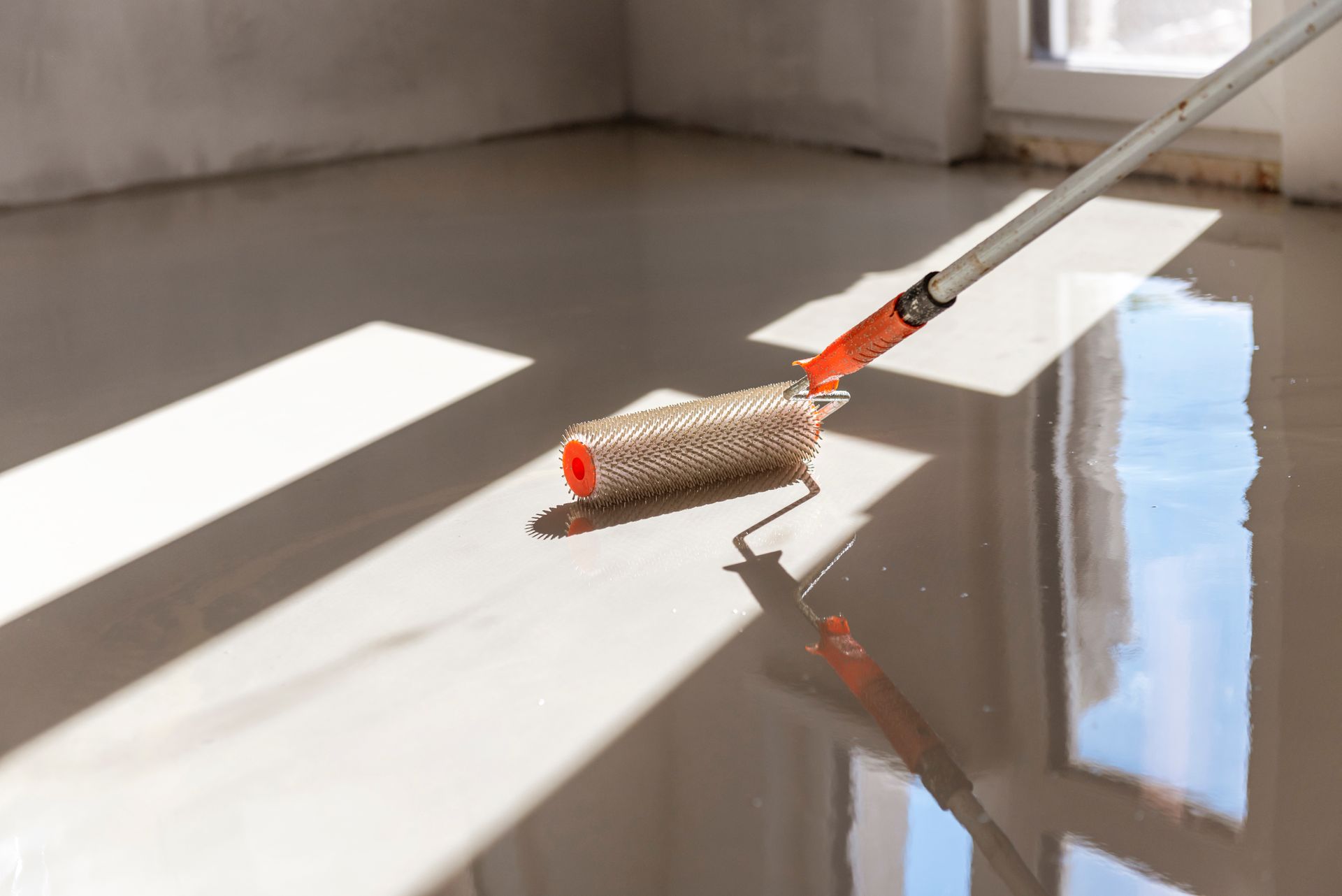 Roller applying glossy, light-colored sealant to a smooth floor in a room with window light.