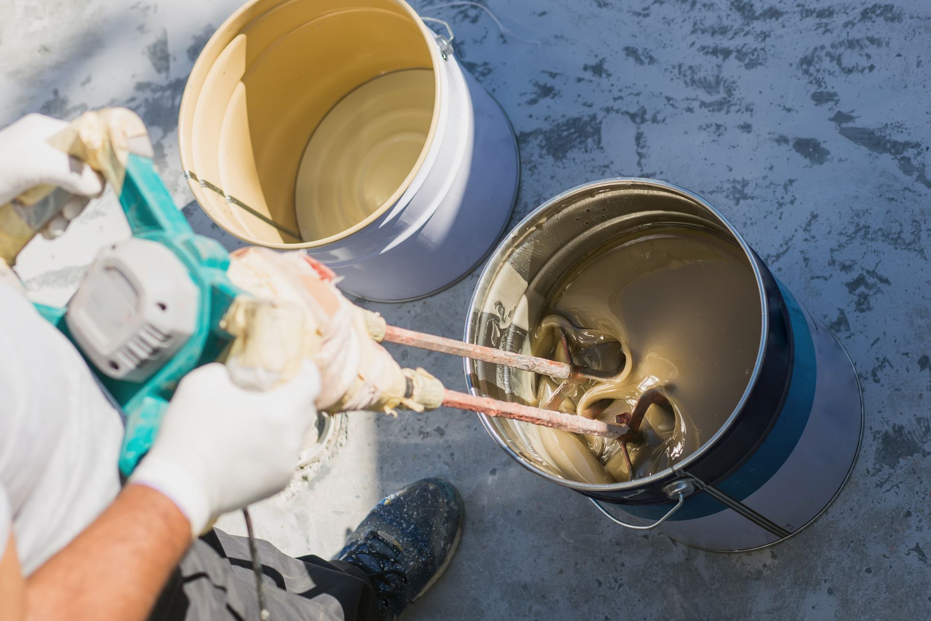 Person mixing a thick, light-brown substance in a bucket with a power mixer. Another bucket is visible.